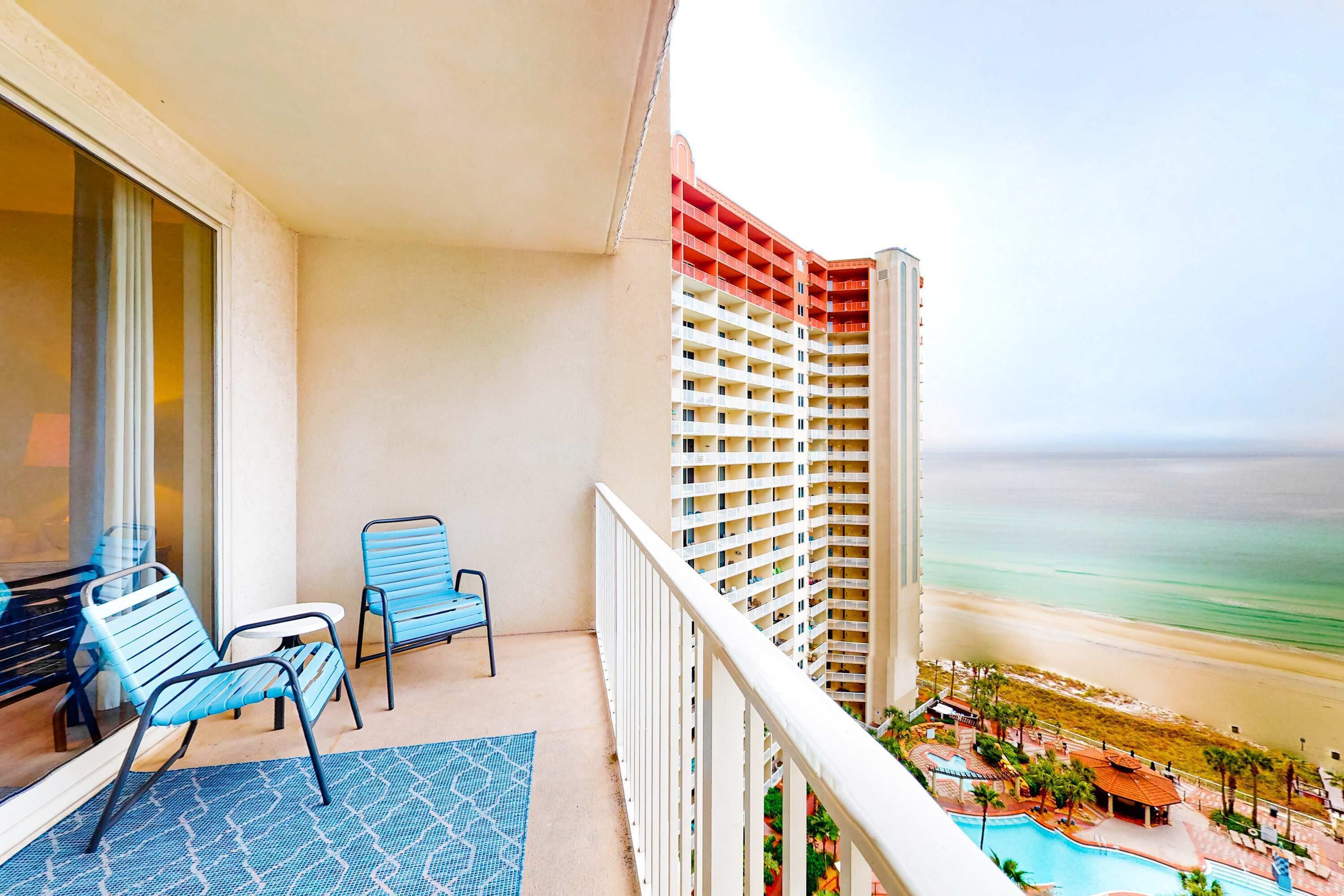 Ein Balkon mit zwei Stühlen und einem gemusterten Teppich, der Blick auf einen Strand und ein Hochhaus mit Poolbereich freigibt.