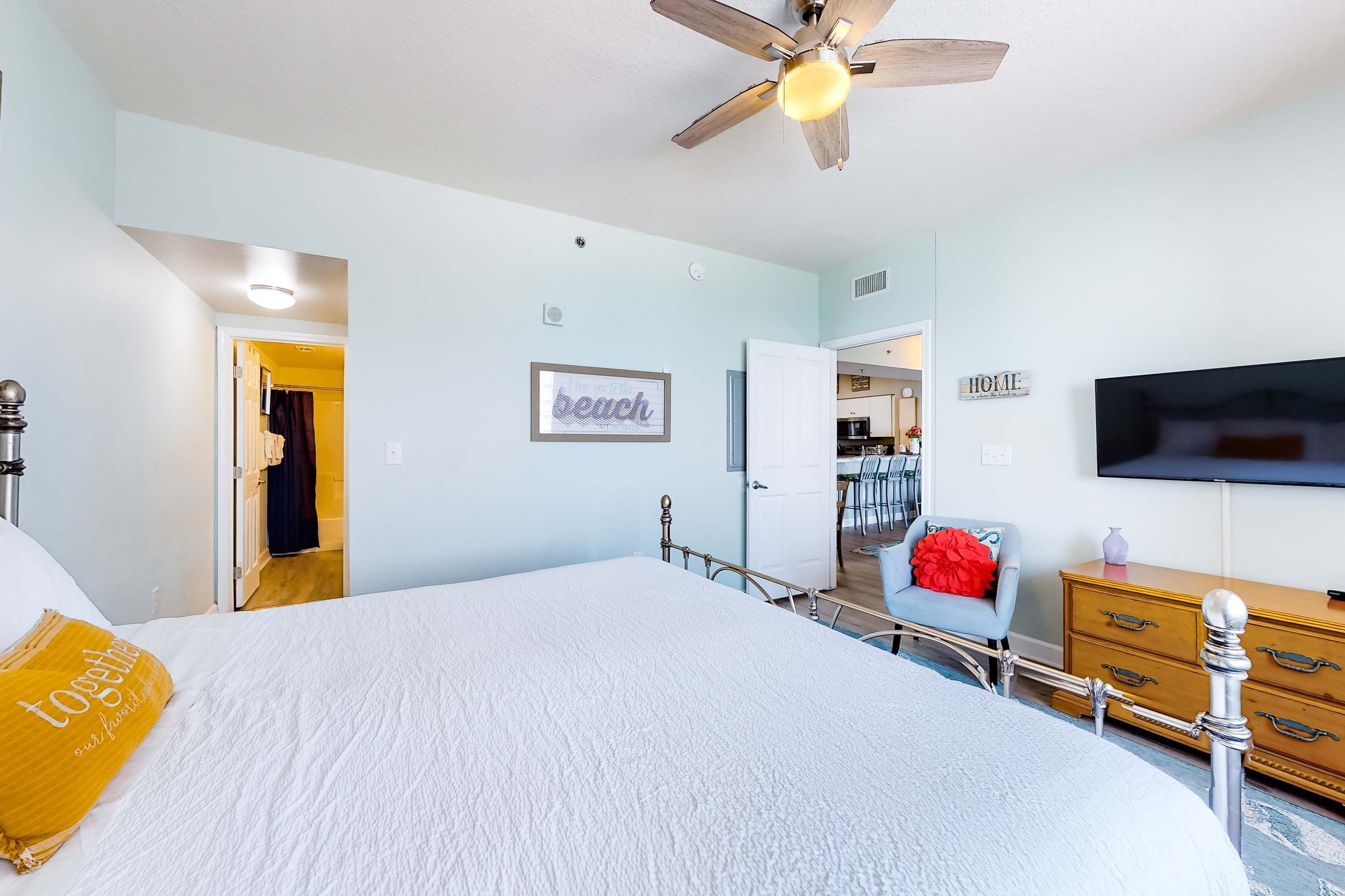 A bedroom with a large bed, a ceiling fan, a TV, and a door leading to a bathroom.