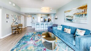 Un salón espacioso con un sofá azul, una mesa de centro y una alfombra colorida. Se ve una cocina al fondo con una barra y sillas.