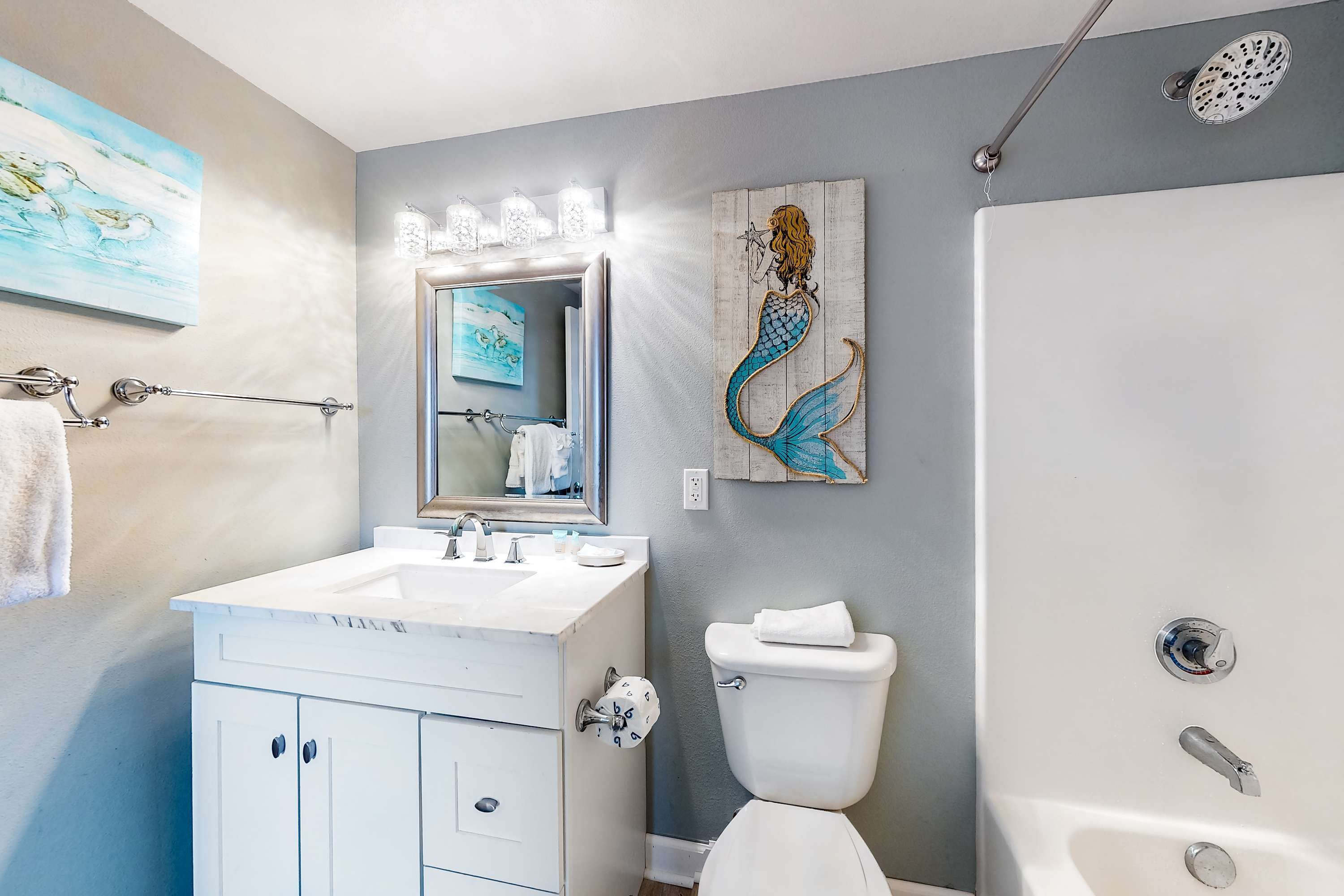 A bathroom with a white sink, a mirror, a toilet, and a shower. There are wall-mounted lights above the mirror and a mermaid painting on the wall.