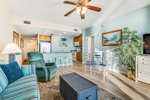 A modern living room with a sofa, armchair, and coffee table. There is a dining area with a table and chairs. The kitchen is visible in the background with cabinets and appliances. - Snow-bird friendly condo with pool & fitness room - walk right out to the sand (Panama City Beach)