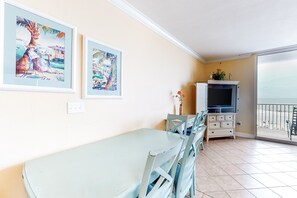 A dining area with a table and chairs, a television on a cabinet, and a balcony with a view. - Charming, waterfront condo with pools, fitness center & beach access (Panama City Beach)