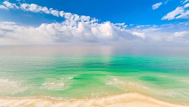 En strand med klart turkis vand og en sandet kystlinje under en blå himmel med spredte skyer.