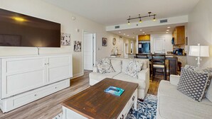A spacious living room with a large white cabinet, a wooden coffee table, and two sofas with patterned cushions. A kitchen area is visible in the background with wooden cabinets and a dining table. - Updated beachfront retreat with hot tub, outdoor pool & balcony (Panama City Beach)