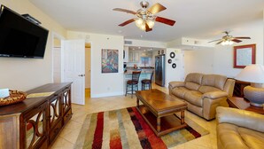A spacious living room with a large sofa, a coffee table, and a TV mounted on the wall. There are two ceiling fans and a kitchen area visible in the background. - Beautiful beachfront getaway with balcony, pools, tennis, and WiFi (Panama City Beach)