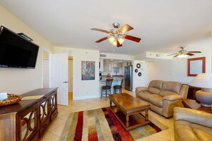 A spacious living room with a large sofa, a coffee table, and a TV mounted on the wall. There are two ceiling fans and a kitchen area visible in the background. - Beautiful beachfront getaway with balcony, pools, tennis, and WiFi (Panama City Beach)