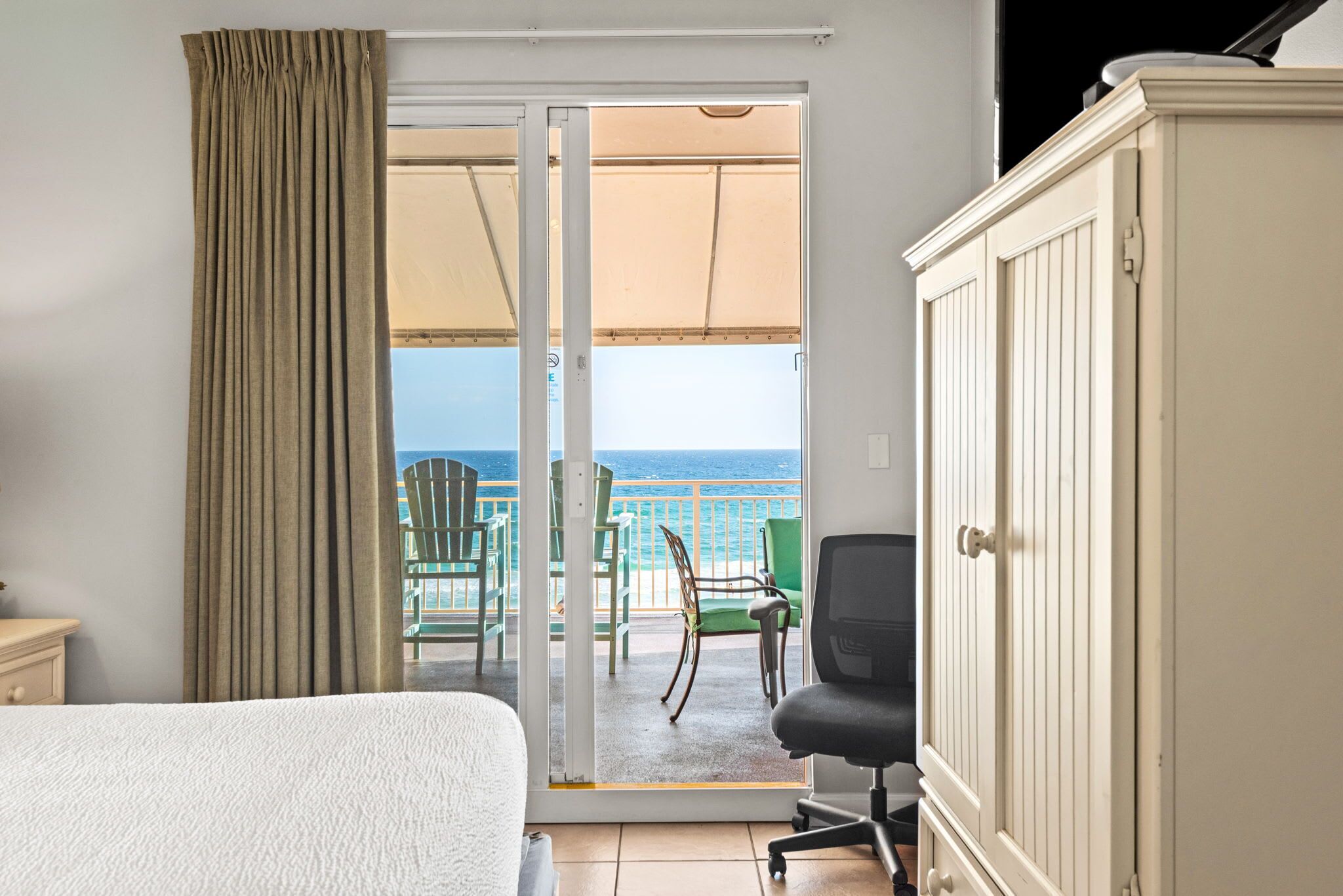 Habitación de hotel con cama, escritorio, silla, armario y puerta corrediza de vidrio que da a un balcón con sillas y vista al océano.