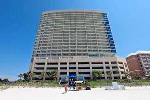 A multi-story hotel with a curved facade, a parking garage below, and a beachside location with palm trees. - Nautical getaway just steps from the beach with pool & hot tub (Panama City Beach)