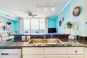 A kitchen with a granite countertop, stainless steel sink, and white cabinets. There is a ceiling fan, a window with curtains, and a TV mounted on the wall. - Beach views from the private balcony at spacious Tidewater condominium (Panama City Beach)