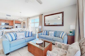A modern living room with two light blue sofas, a glass-top coffee table, and a kitchen in the background. - Oceanfront, 7th-floor condo with pool, ocean view & high-speed WiFi (Panama City Beach)