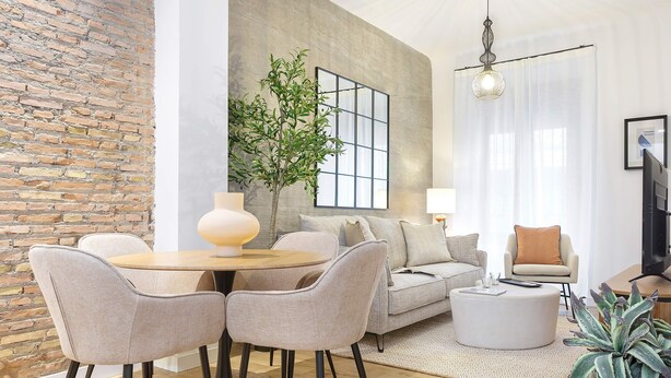 Un salón moderno con una mesa redonda, dos sillas, un sofá, una mesa auxiliar, una planta en maceta y un amplio ventanal con vistas.