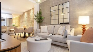 Una sala moderna con sofá, reposapiés y sillas, un comedor con mesa y sillas, y una cocina al fondo.