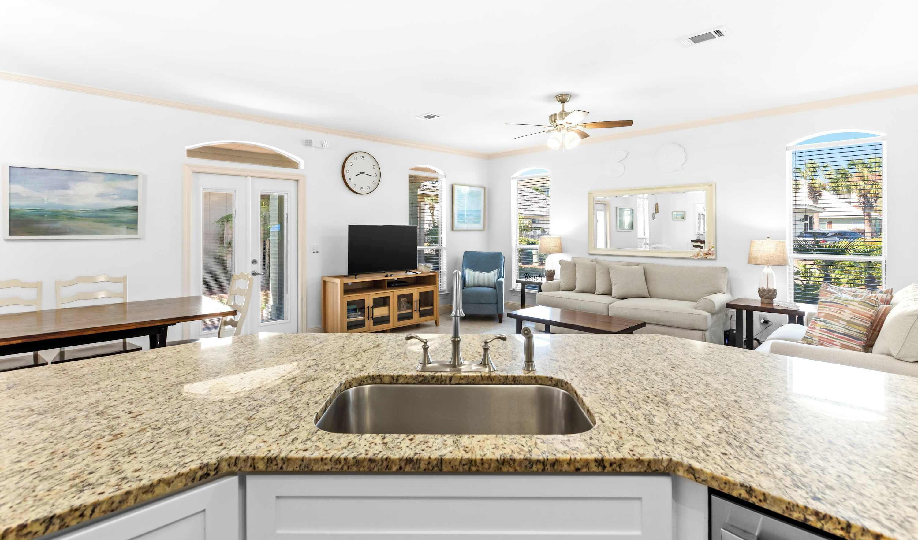 A kitchen with granite countertops, a stainless steel sink, a dining area with a wooden table and chairs, a living room with a sofa and a TV, and a large window with blinds.