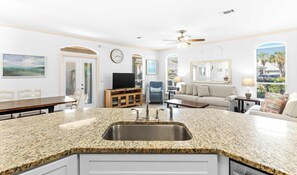 A kitchen with granite countertops, a stainless steel sink, a dining area with a wooden table and chairs, a living room with a sofa and a TV, and a large window with blinds. - Gorgeous coastal private home, close to everything and shared pool (Miramar Beach)