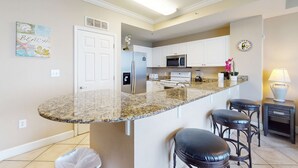 A kitchen with a granite countertop, white cabinets, a stainless steel refrigerator, a microwave, and a sink. There are three bar stools and a small table with a lamp. - Beach-chic gulf-front condo w resort pools, hot tubs & beach service (Panama City Beach)