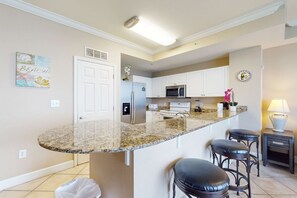 Eine Küche mit Granit Arbeitsplatte, weißen Schränken, einem Edelstahl-Kühlschrank, einer Mikrowelle und einem Spülbecken. Drei Barhocker und ein kleiner Tisch mit Lampe ergänzen das Bild.