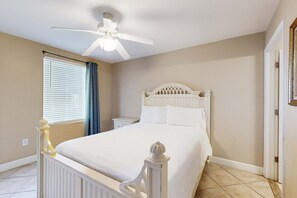 Une chambre à coucher avec un lit blanc, un ventilateur de plafond, une fenêtre avec des stores et une porte.