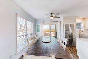 A dining area with a wooden table and chairs, a ceiling fan, and a kitchen with a stainless steel refrigerator. - Ideally located coastal condo with Gulf views, private balcony & beach access (Perdido Key)