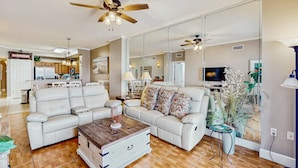 A spacious living room with a large sofa, a coffee table, and a TV. There is a kitchen area in the background with cabinets and a refrigerator. - Gorgeous beachfront condo, shared pool and beach access (Panama City Beach)