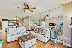 A spacious living room with a large sofa, a coffee table, and a TV. There is a kitchen area in the background with cabinets and a refrigerator. - Gorgeous beachfront condo, shared pool and beach access (Panama City Beach)
