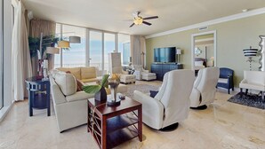 A spacious living room with a large window, a ceiling fan, and a flat-screen TV. - Elegant, Gulf-Front Condo w/ Large Balcony, Beach Access & Shared Pool/Gym (Perdido Key)