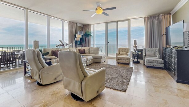 A spacious living room with a large window overlooking the ocean, a ceiling fan, and a television. - Elegant, Gulf-Front Condo w/ Large Balcony, Beach Access & Shared Pool/Gym (Perdido Key)