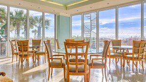 A spacious dining area with a high ceiling fan, large windows with a view of palm trees, and a spiral staircase. - Elegant, Gulf-Front Condo w/ Large Balcony, Beach Access & Shared Pool/Gym (Perdido Key)