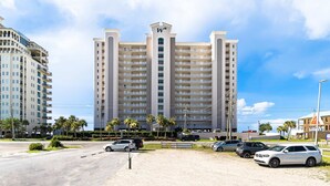 A multi-story hotel with a central tower, a parking lot with several cars, and palm trees in the foreground. - Inviting Gulf-front condo In Perdido Key with waterfront pool/hot tub and oceanfront balcony (Perdido Key)