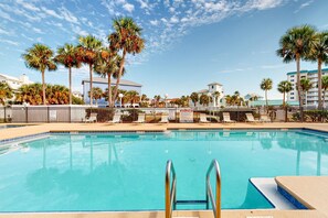 A swimming pool with a metal ladder, surrounded by palm trees and lounge chairs, with buildings in the background. - Sunny condo with pool, hot tub, pool views, balcony, & AC (Perdido Key)