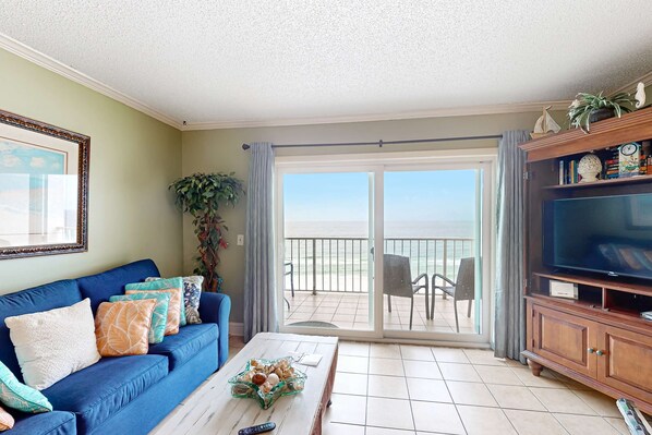 A living room with a blue sofa, a coffee table with decorative items, a television on a wooden cabinet, and a sliding glass door leading to an outdoor balcony with chairs. - Oceanfront condo with beach access, pools & hot tubs (Panama City Beach)