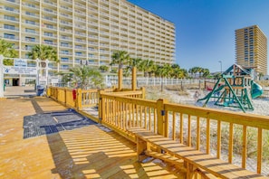 A wooden boardwalk leading to a beach area with a playground and a large hotel building in the background. - Oceanfront condo with beach access, pools & hot tubs (Panama City Beach)