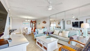 Ein geräumiges Wohnzimmer mit einem weißen Couchtisch, einer Couch und einem Fernseher. Ein Deckenventilator und ein großer Spiegel spiegeln den Raum wider.