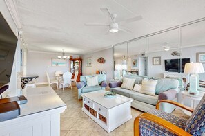 A spacious living room with a white coffee table, a sofa, and a TV. There is a ceiling fan and a large mirror reflecting the room. - Beachfront condo with beach setup- close to everything (Fort Walton Beach)