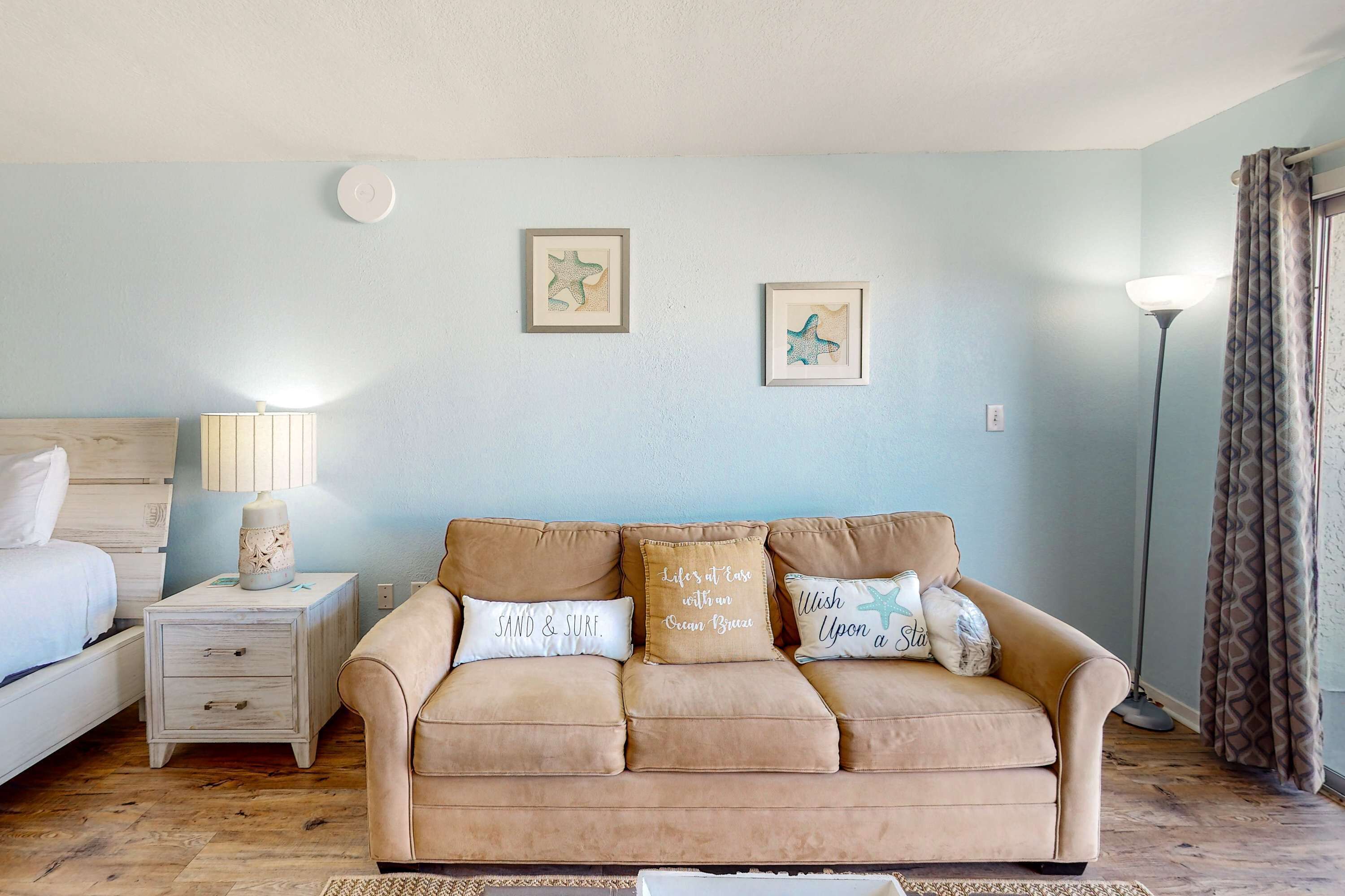 A bedroom with a beige sofa, a bedside table with a lamp, and two framed pictures on the wall.