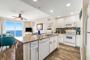 A modern kitchen with white cabinets, a granite countertop, and a large island. There is a ceiling fan, a microwave, and a refrigerator. - Beachfront condo with amazing balcony views, pool & complimentary beach chairs (Panama City Beach)