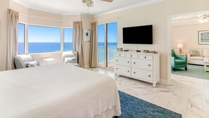 Une chambre d’hôtel avec un grand lit, un ventilateur de plafond, une télévision, un balcon avec vue sur l’océan et une commode blanche.