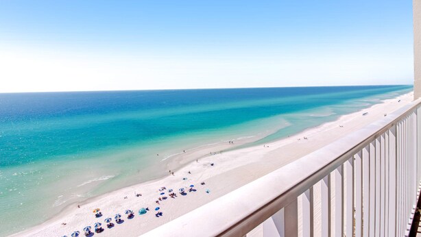 Une vue sur la plage depuis un balcon doté d’une balustrade, avec une mer d’un bleu limpide et du sable blanc.