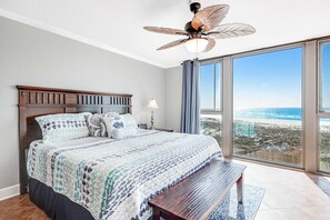 A bedroom with a large bed, a wooden headboard, a ceiling fan, a nightstand, a lamp, and a view of the ocean through large windows. - Gulf-front condo with pool & tennis court access, W/D, & AC (Perdido Key)