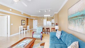 Una sala de estar con un sofá azul, una mesa de centro de madera y sillas con estampado. Se aprecia una cocina al fondo con armarios blancos y una zona de bar.