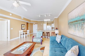 A living room with a blue sofa, a wooden coffee table, and patterned chairs. There is a kitchen in the background with white cabinets and a bar area. - Picturesque beachfront condo with gym, BBQ area, pools & hot tub (Panama City Beach)