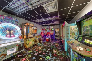 Een helder verlichte speelhal met diverse spelautomaten, een vloer met een stervenspatroon en een plafond met een geometrisch ontwerp.