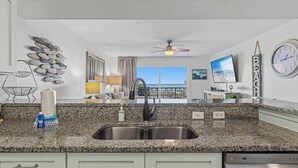 A kitchen with granite countertops, a stainless steel sink, a ceiling fan, and a view of the ocean through a large window. - Oceanfront Condo with Shared Pool & Hot Tub, Ocean View, & Private Washer&Dryer (Fort Walton Beach)