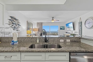 A kitchen with granite countertops, a stainless steel sink, a ceiling fan, and a view of the ocean through a large window. - Oceanfront Condo with Shared Pool & Hot Tub, Ocean View, & Private Washer&Dryer (Fort Walton Beach)
