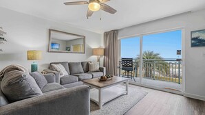 A modern living room with a ceiling fan, a sofa, a coffee table, and a view of the outdoors through a sliding glass door. - Oceanfront Condo with Shared Pool & Hot Tub, Ocean View, & Private Washer&Dryer (Fort Walton Beach)