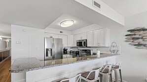 A modern kitchen with white cabinets, a granite countertop, a stainless steel refrigerator, a microwave, and a coffee maker. There are three bar stools and a fish wall decoration. - Oceanfront Condo with Shared Pool & Hot Tub, Ocean View, & Private Washer&Dryer (Fort Walton Beach)