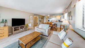 A modern living room with a flat-screen TV, a wooden coffee table, a sofa, and a dining area with a table and chairs. - Luxury, oceanfront condo with outdoor pool & patio - snowbird-friendly (Miramar Beach)