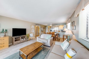A modern living room with a flat-screen TV, a wooden coffee table, a sofa, and a dining area with a table and chairs. - Luxury, oceanfront condo with outdoor pool & patio - snowbird-friendly (Miramar Beach)
