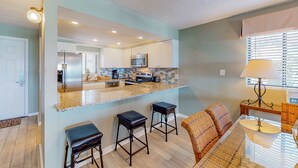 A modern kitchen with a central island, bar stools, and a dining area with a table and chairs. - Luxury, oceanfront condo with outdoor pool & patio - snowbird-friendly (Miramar Beach)