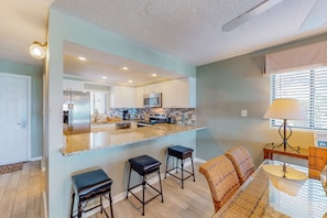 A modern kitchen with a central island, bar stools, and a dining area with a table and chairs. - Luxury, oceanfront condo with outdoor pool & patio - snowbird-friendly (Miramar Beach)