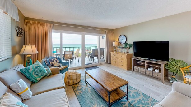A spacious living room with a sofa set, a coffee table, a TV, and a large window offering a view of the ocean. - Luxury, oceanfront condo with outdoor pool & patio - snowbird-friendly (Miramar Beach)